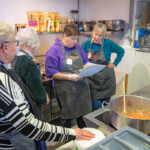 The group in charge of cooking the soup in the kitchen. Two of the members are looking over the recipe while the others await further instructions. There's a big metal pot on the hob with shopped vegetables in.