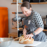 A member of the group smiling as she holds a loaf of bread down with one hand and cuts it into slices with the other.