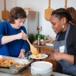 A quiche being served from a metal tray onto a plate held by Wanja,