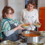 Jane, the tutor, is serving up bowls of soup made by the group. She's using a stainless steel ladel to scoop it up out of the metal pot. Clare is standing by her side with a bowl in one hand, waiting for another bowl to take to the dining table.