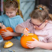Two young girls sat side by side, both using spoons to remove the innards from their pumpkins.