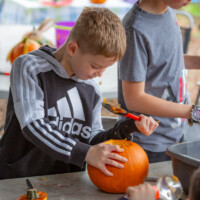 A young boy standing at the table, one hand holding his pumpkin still while the other uses a scoot to remove the innards.