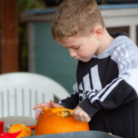 A young boy standing up and using a spoon to remove innards from a pumpkin.