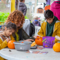 Two kids, both in yellow jackets, sat at a table. The girl on the left is drawing a design on her pumpkin. The boy on the right is waiting for Katie, also sat down, to finish carving his pumpkin.