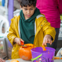 A young boy in a yellow jacket holding a scoop in one hand and using the other hand to deposit some pumpkin innards into a bucket.