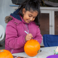 A child smiling as she holds her pumpkin still and draws the design on it.