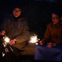 Two children sat on a bench, watching their lit sparklers.