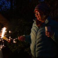 Polly smiling while holding a hot drink in one hand and a lit sparkler in the other.